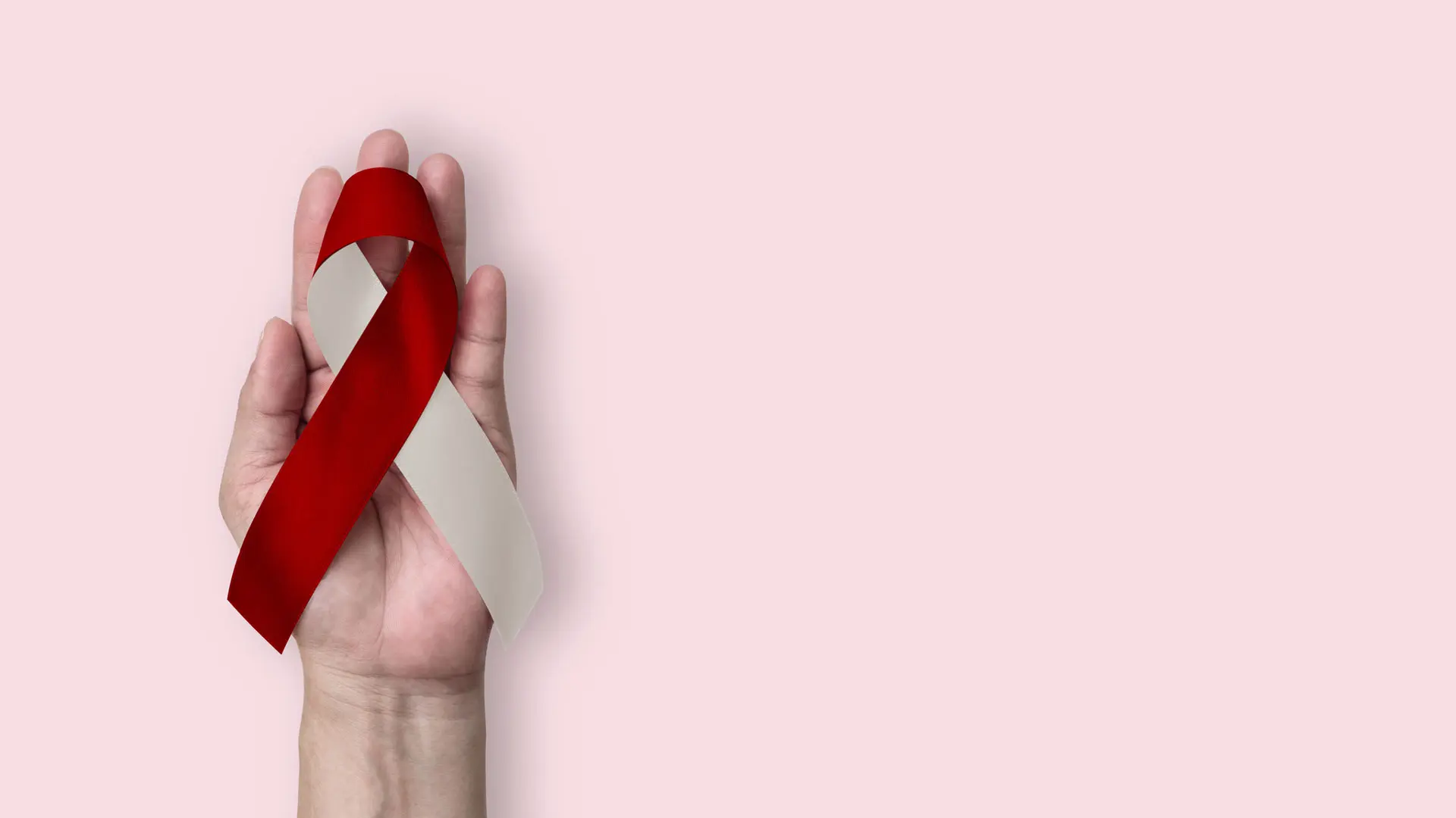 Photograph of a hand holding a red and white oral cancer awareness ribbon against a light pink background