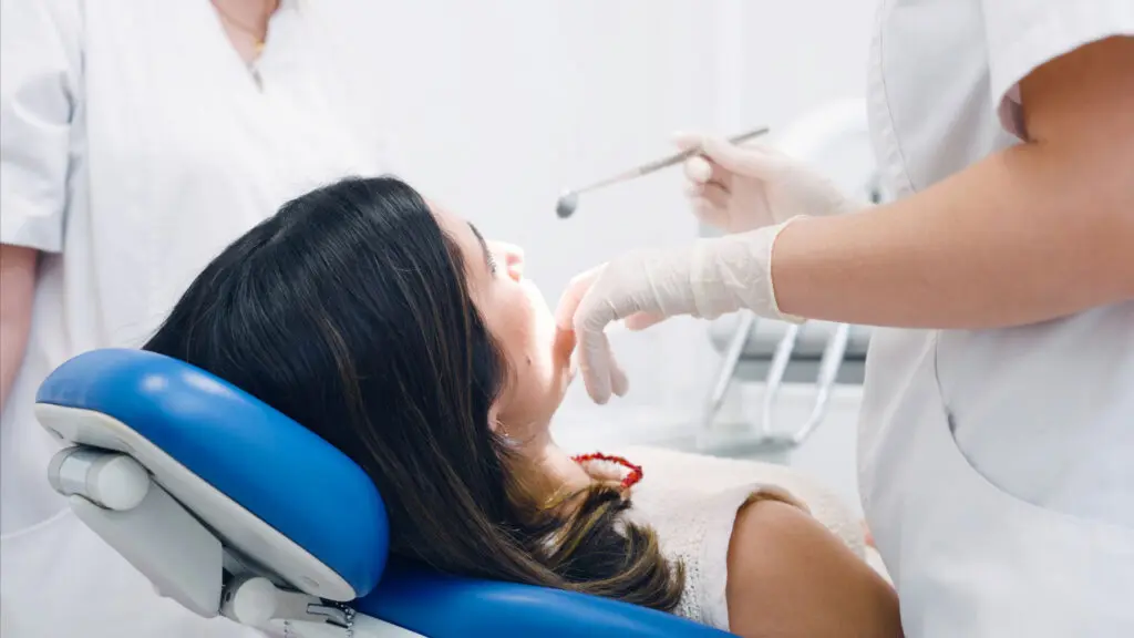 Woman with medium length brown hair sits in a dentist chair facing away from the camera towards two dental professionals in white scrubs who are about to perform an oral exam