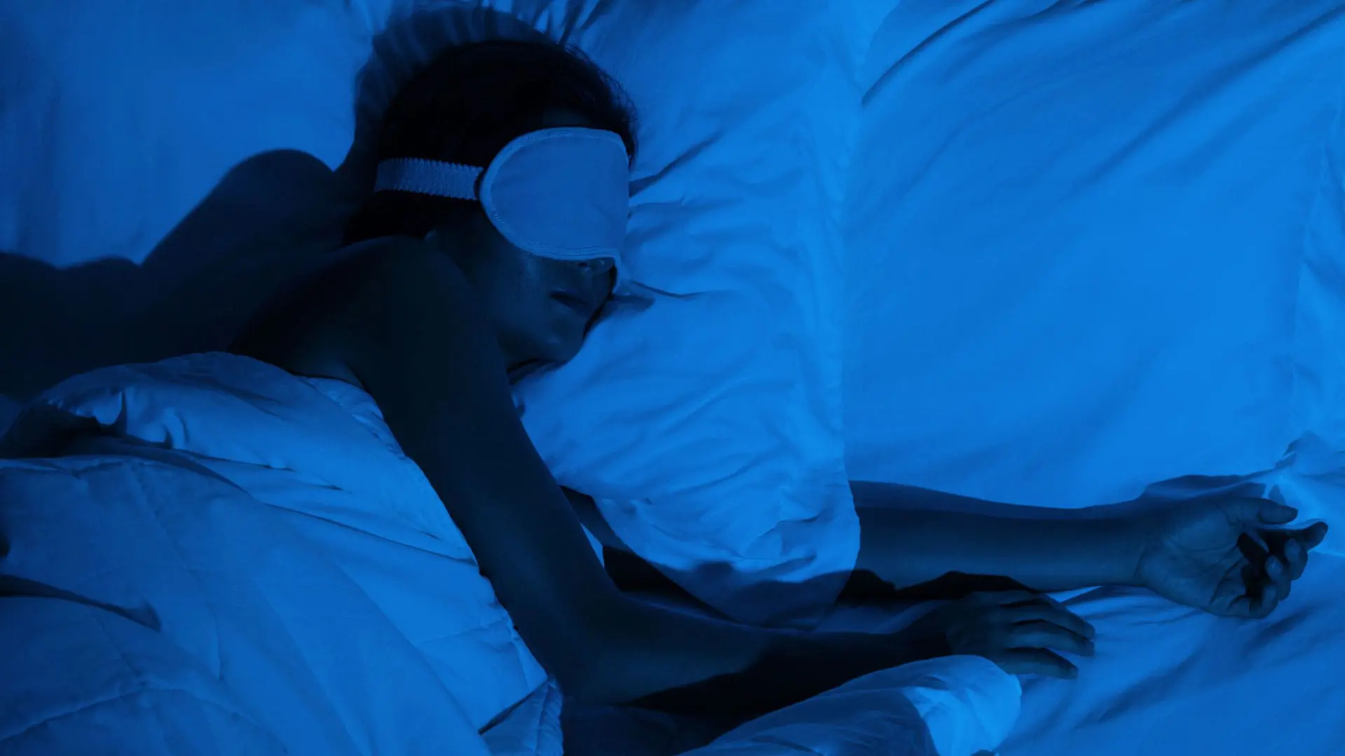 A woman in a sleep mask peacefully sleeps on her side in a dark room.